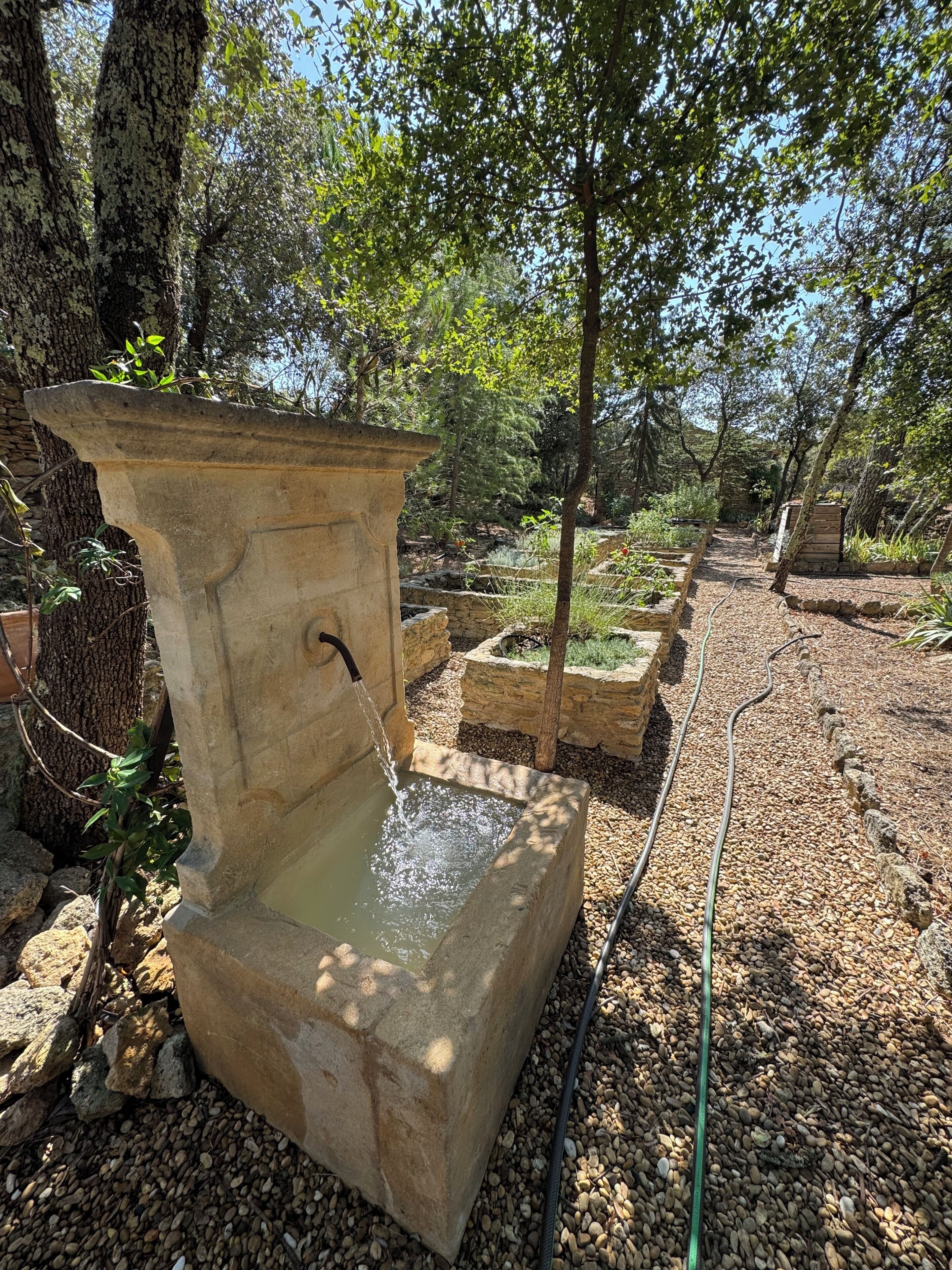 A stone water feature with a spout, surrounded by trees and garden beds.