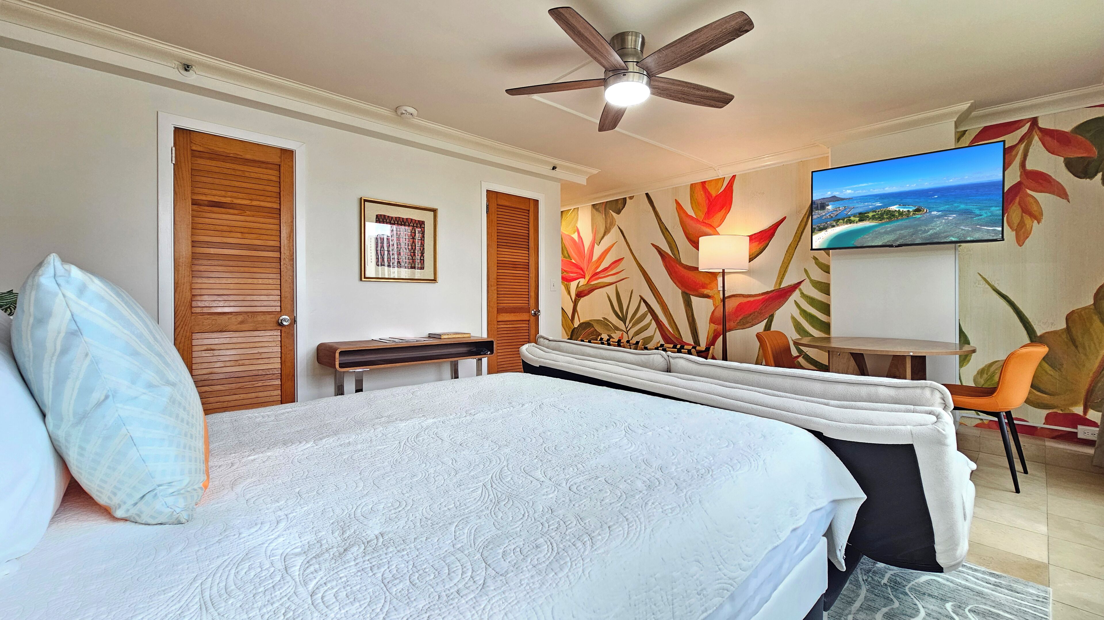 Une chambre d’hôtel équipée d’un lit, d’un bureau, d’une chaise, d’un ventilateur de plafond et d’un téléviseur fixé au mur.