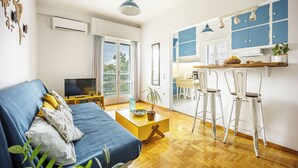 A modern living room with a sofa, a coffee table, and a TV. There is a kitchen area with blue cabinets and white chairs. - Amazing Acropolis View From A Sunny Renovated Apt in Athens! (Athina)