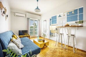 A modern living room with a sofa, a coffee table, and a TV. There is a kitchen area with blue cabinets and white chairs. - Amazing Acropolis View From A Sunny Renovated Apt in Athens! (Athina)