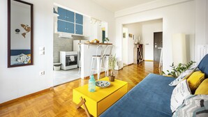 A modern living room with a yellow coffee table, a blue sofa, and a kitchen area with white cabinets and a blue accent wall. - Amazing Acropolis View From A Sunny Renovated Apt in Athens! (Athina)