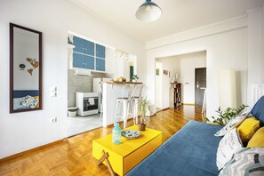 A modern living room with a yellow coffee table, a blue sofa, and a kitchen area with white cabinets and a blue accent wall. - Amazing Acropolis View From A Sunny Renovated Apt in Athens! (Athina)