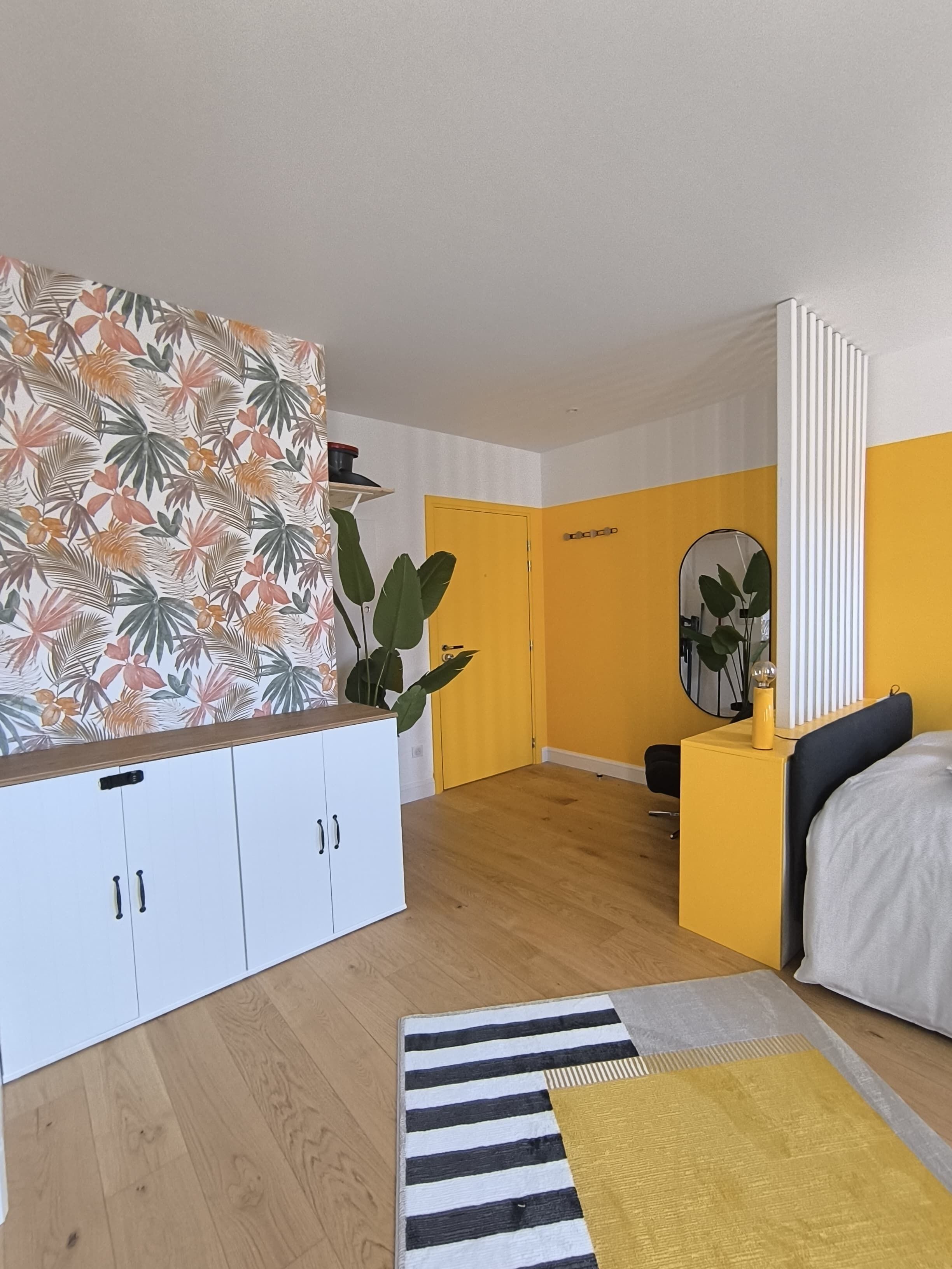 A bedroom with a bed, a bedside table, a mirror, a wardrobe, and a large plant.