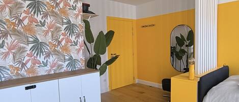A bedroom with a bed, a bedside table, a mirror, a wardrobe, and a large plant.