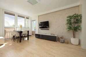 A modern living room with a dining area, a flat-screen TV mounted on a brick wall, a large potted plant, and a sliding glass door leading to an outdoor space. - Family Apartment | Workplace, Close to Metro (Warszawa)