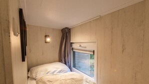 Ein kleines Zimmer mit einem Einzelbett, einem Fenster, einer Wandleuchte und einer Tür.