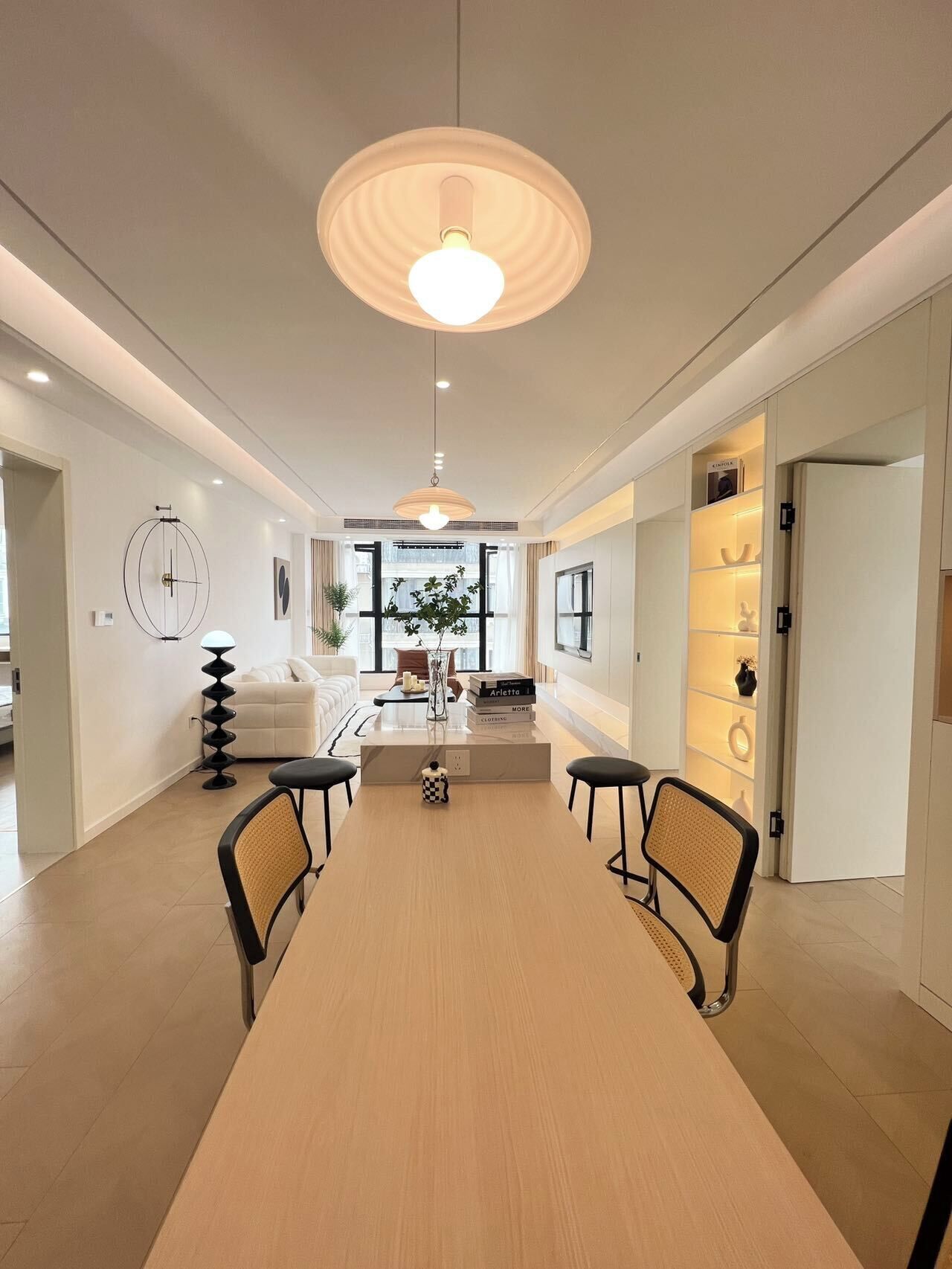 A modern dining area with a long table, two chairs, a hanging light fixture, a sofa, a coffee table, and a bookshelf.