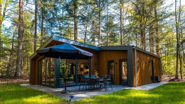 Una cabaña moderna con cerramiento de vidrio, mobiliario exterior y un bosque de fondo.