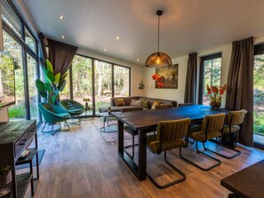 A modern dining area with a large table, chairs, and a view of the forest through large windows. - Vakantiepark De Wiltzangh - Zandborg Lodge by Interhome (Ruinen)