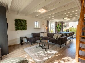A modern living room with a sectional sofa, a coffee table, a small side table, a large green wall decoration, a dining area with a table and chairs, and a wooden ladder. - Vakantiepark Wiedense meren - Weerribben by Interhome (Wanneperveen)