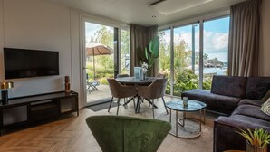 Een moderne woonkamer met een flatscreen televisie, een groene fauteuil, een donkere bank, een eethoek met tafel en stoelen, en grote ramen met uitzicht op het buitenleven.