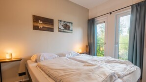 Een slaapkamer met een bed, nachtkastjes, een raam met gordijnen en twee kunstwerken aan de muur.
