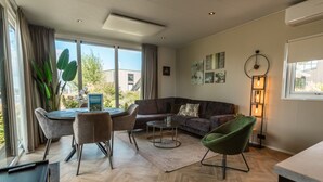 Een moderne woonkamer met een bank, stoelen, een salontafel en een eettafel. Er zijn grote ramen, een vloerlamp en kunst aan de muren.