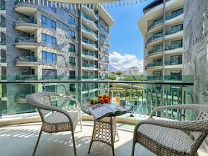 Ein Balkon mit Korbmöbeln, einem Tisch mit Obst und einer Zeitung sowie Blick auf mehrstöckige Gebäude.