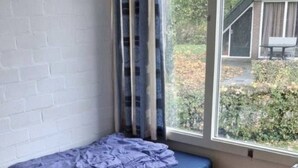 Une petite pièce avec un lit simple, une fenêtre avec des rideaux, un mur en briques et un radiateur.