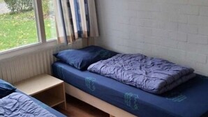 Une chambre avec deux lits simples, une fenêtre avec des rideaux, un tableau encadré au mur et un mur en briques blanches.