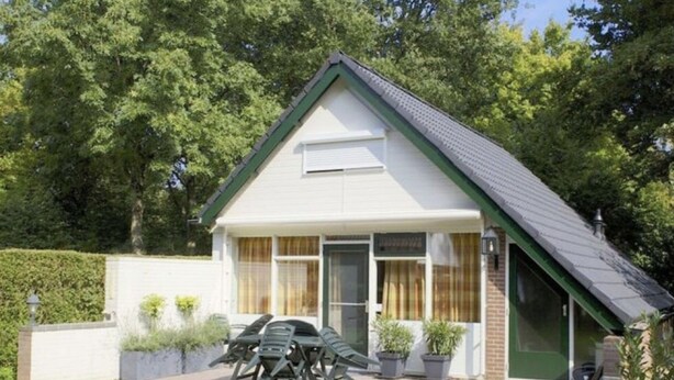 Une maison de plain-pied avec un toit en pente, une véranda à l’avant avec une table et des chaises, et un petit jardin agrémenté de plantes en pot.