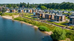 A residential area with modern houses by a river, featuring a mix of single and multi-story buildings, a sandy beach, and green lawns. - Vakantiepark Aan de Maas - Weide Lande 4 personen by Interhome (Kerkdriel)