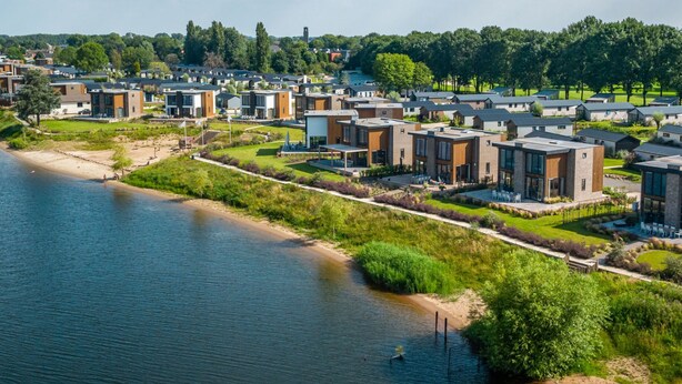 A residential area with modern houses by a river, featuring a mix of single and multi-story buildings, a sandy beach, and green lawns. - Vakantiepark Aan de Maas - Weide Lande 6 personen by Interhome (Kerkdriel)