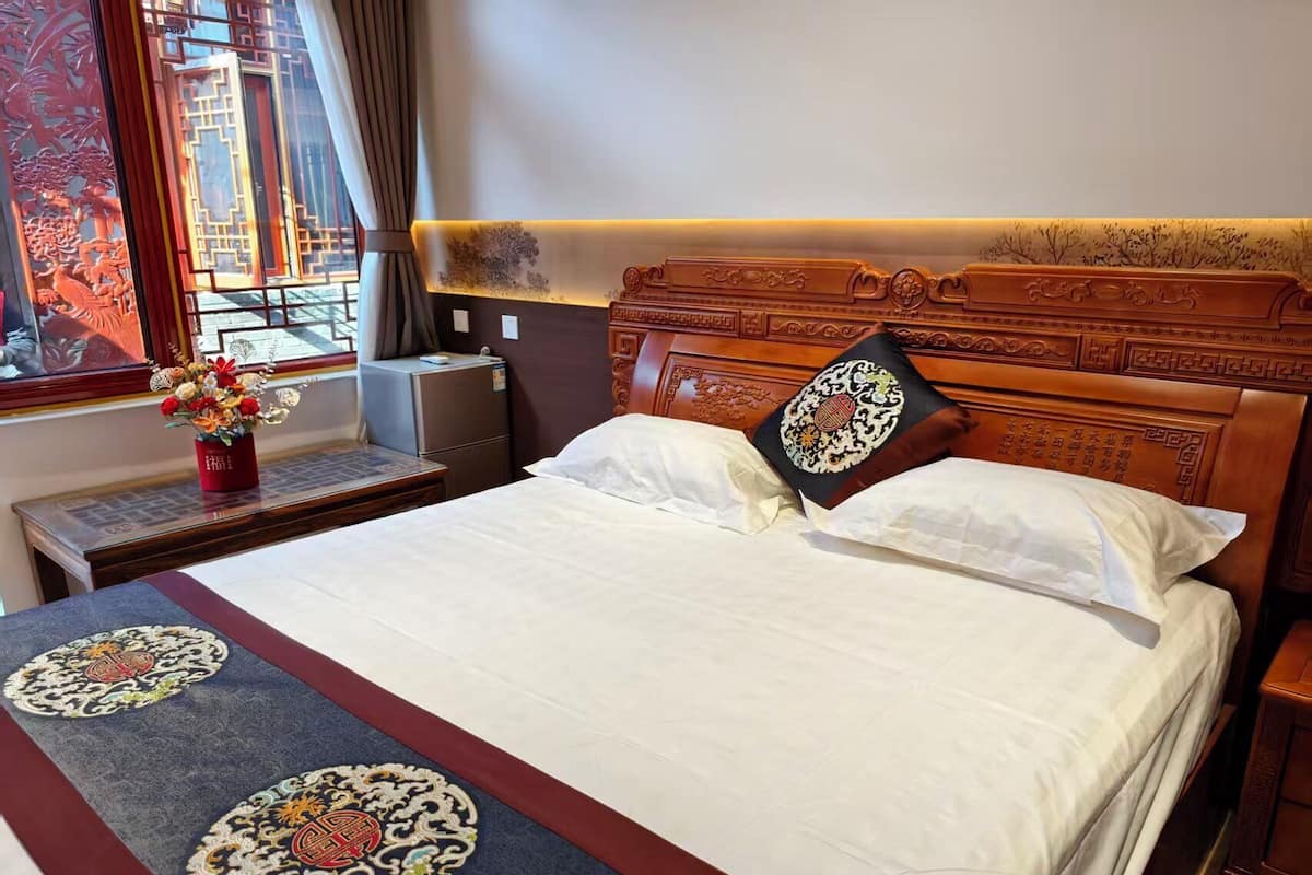 Habitación de hotel con cabecera de madera, cama con ropa blanca, almohada decorativa, mesita de noche con un florero y ventana con vista a edificios.