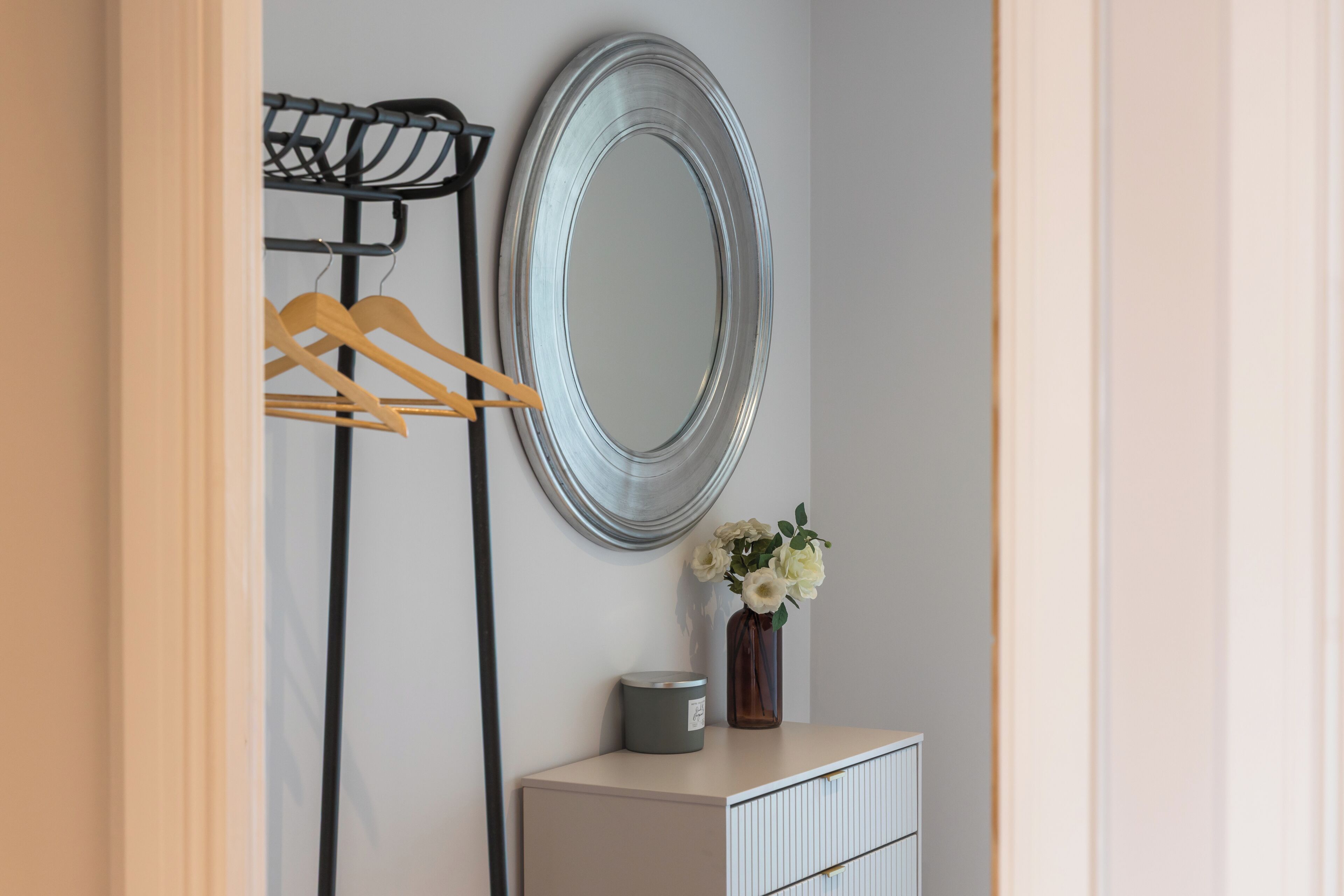 A room with a mirror, a coat rack, a vase with flowers, and a cabinet.