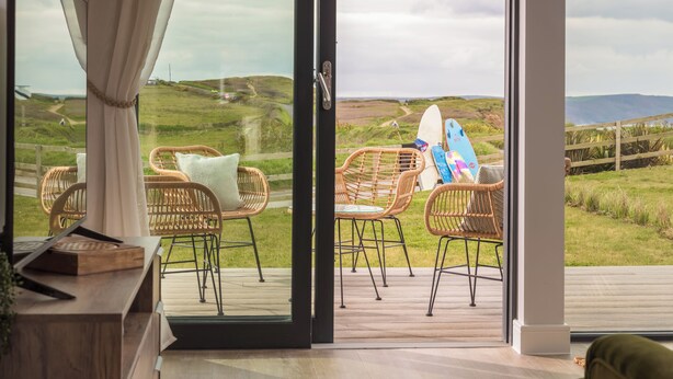 A room with a large glass door leading to an outdoor deck with wicker chairs and surfboards. - Perrantide (Bude)