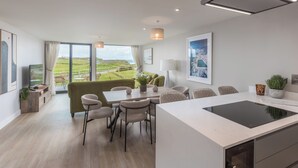 A modern kitchen with a central island, dining area with chairs, and a large window with a view of a green landscape. - Perrantide (Bude)