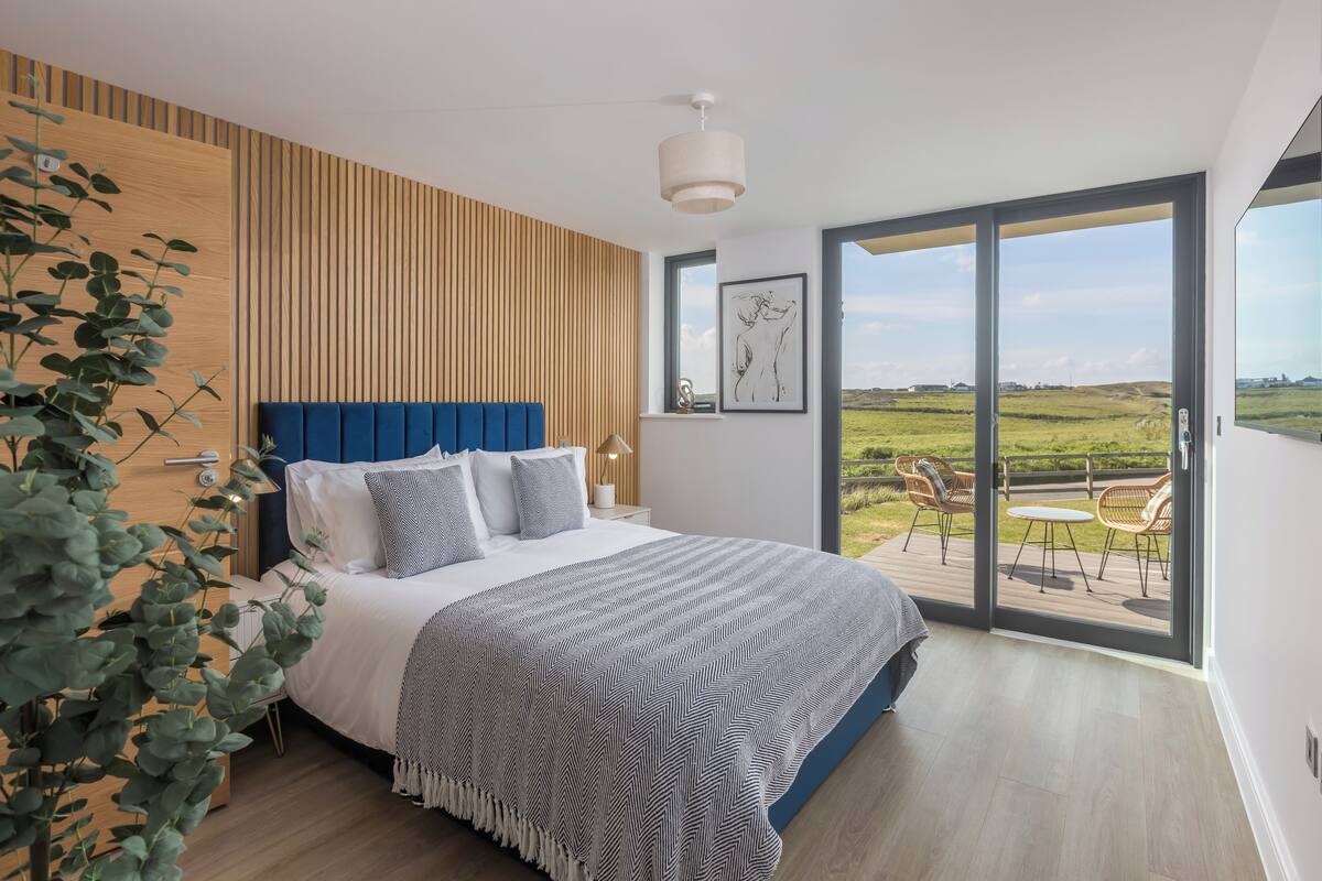 A bedroom with a large bed, a wooden slat wall, a view of the outdoors, and a balcony with chairs.