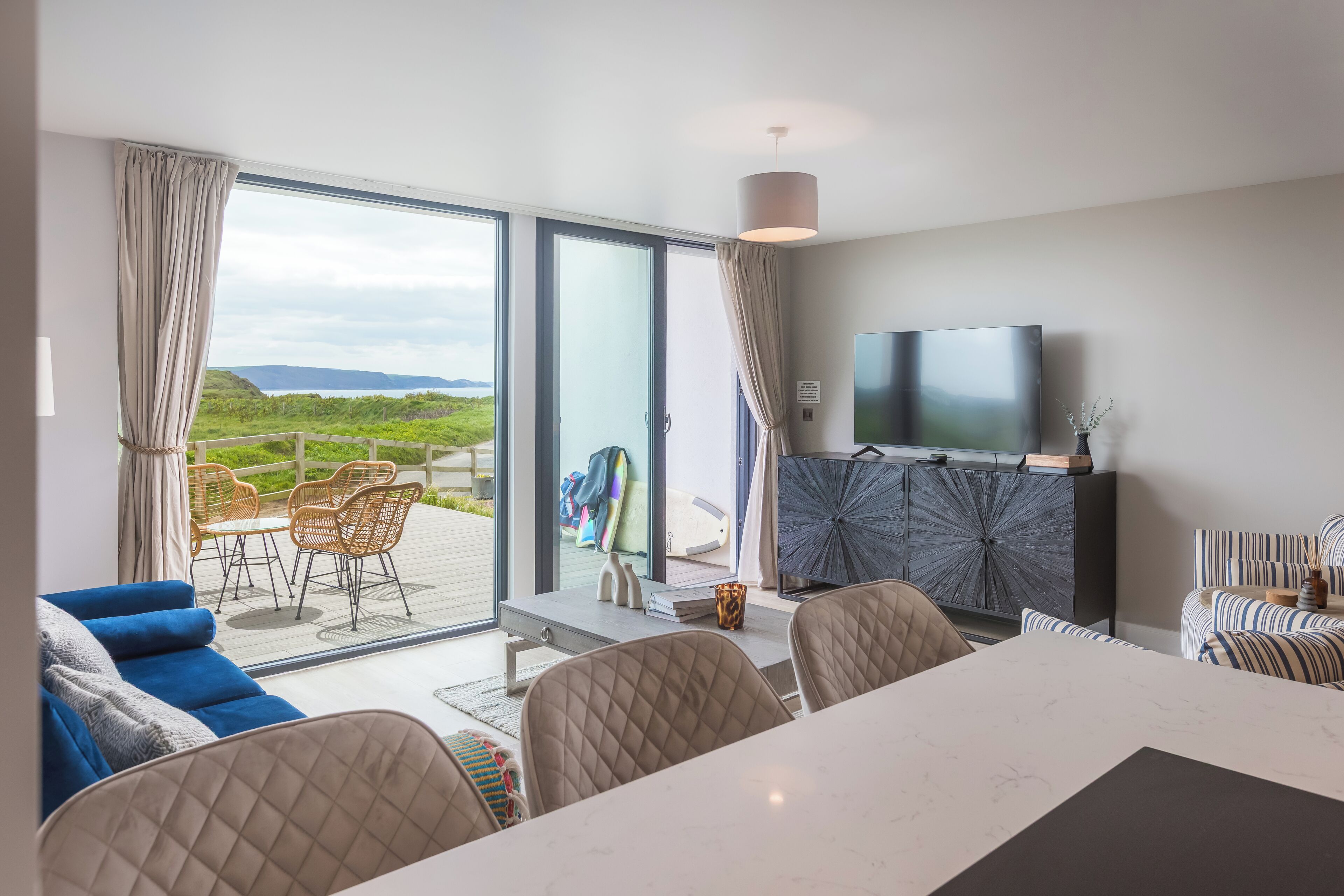A modern living room with a large sliding glass door leading to an outdoor area, a flat-screen TV mounted on the wall, and a dining table with chairs.