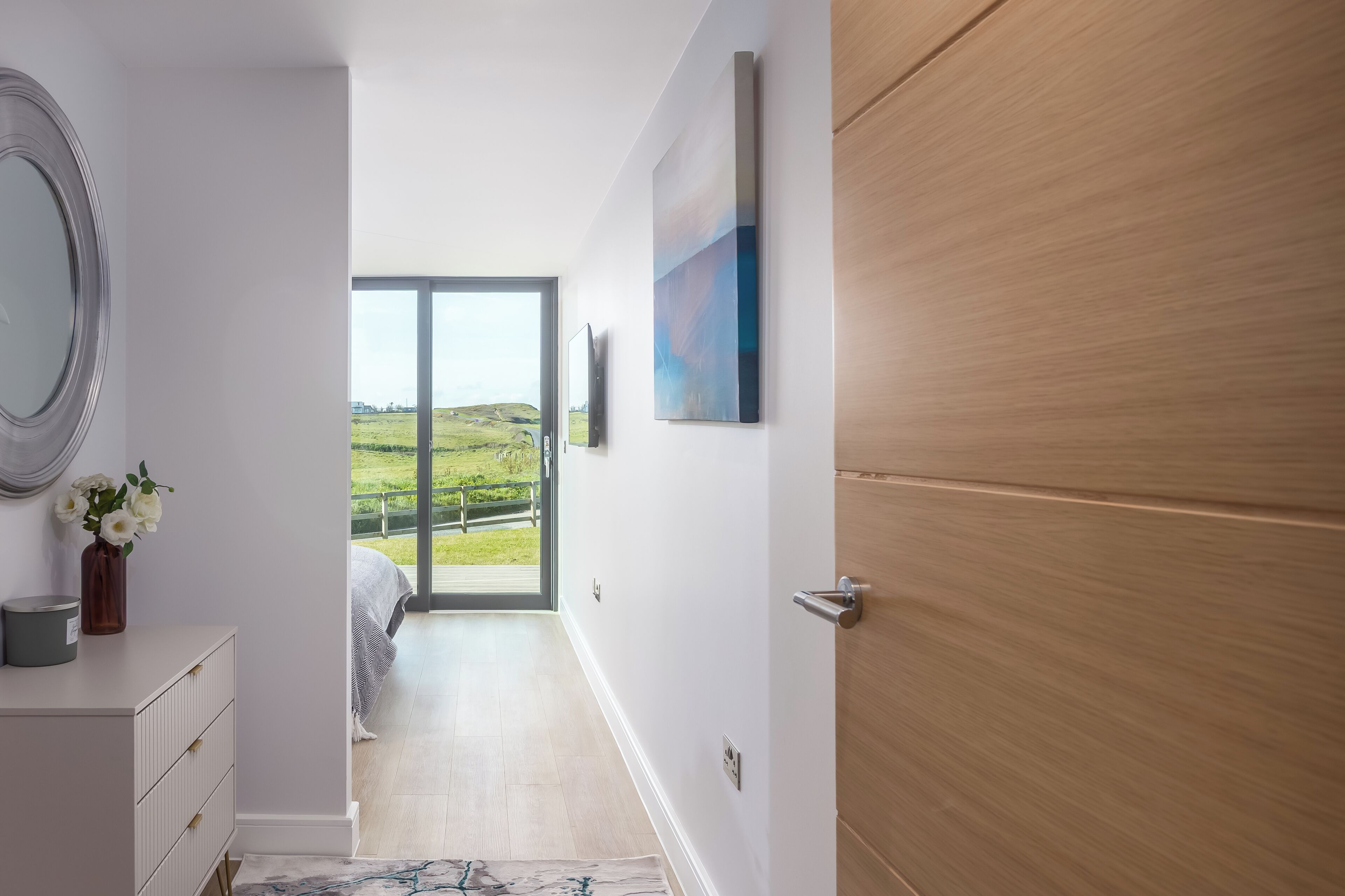 A modern interior with a wooden door, a white wall, a flat-screen TV, and a view of a green landscape through a glass door.