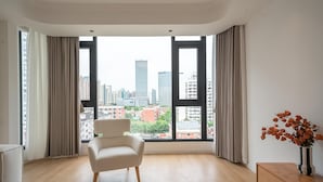 Een moderne woonkamer met een groot raam, een witte fauteuil, een houten dressoir en een vaas met gedroogde bloemen.