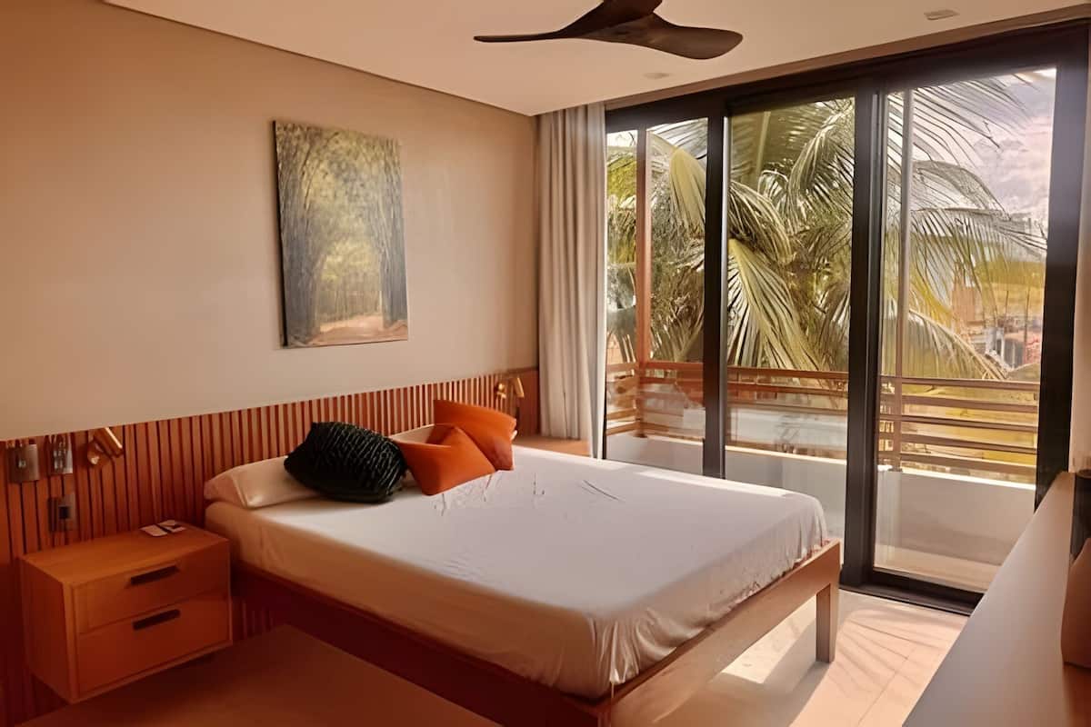 A modern bedroom with a large bed, a nightstand, a ceiling fan, and a view of palm trees through a sliding glass door.