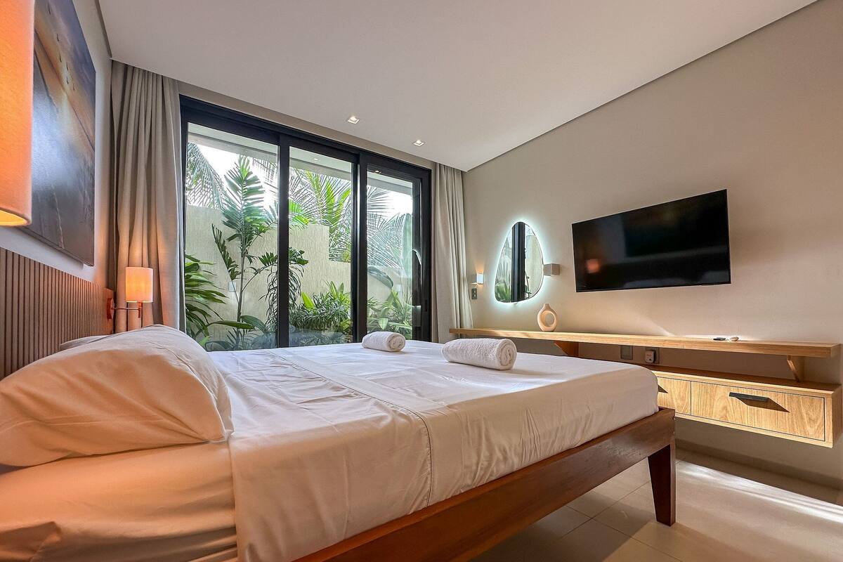 A modern hotel room with a large bed, a TV mounted on the wall, and a view of greenery through the window.