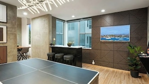 A ping pong table in a room with a wooden floor, a bar area with stools, a large window, and a wall-mounted picture. - Walk to Gaslamp & Petco • Rooftop Spa & Game Room (San Diego)