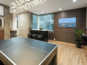 Una mesa de ping pong en una habitación con piso de madera, una zona de bar con taburetes, un ventanal grande y un cuadro colgado en la pared.