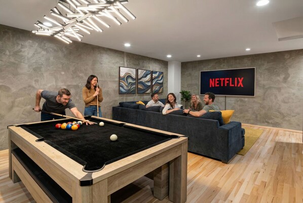 Uma mesa de bilhar em uma sala moderna com uma TV grande exibindo 'NETFLIX' e arte abstrata nas paredes.