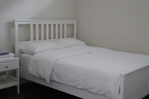 A white single bed with a wooden headboard, a nightstand with a drawer, and a plain wall. - Spacious 2 Basement Bedrooms Near UBC (Vancouver)