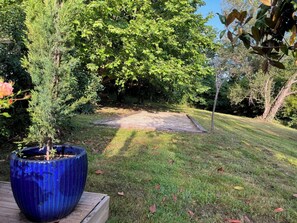 Ein Gartenbereich mit einer Holzterrasse, einem blauen Pflanzgefäß mit einem kleinen Baum und einer Rasenfläche mit Bäumen im Hintergrund.