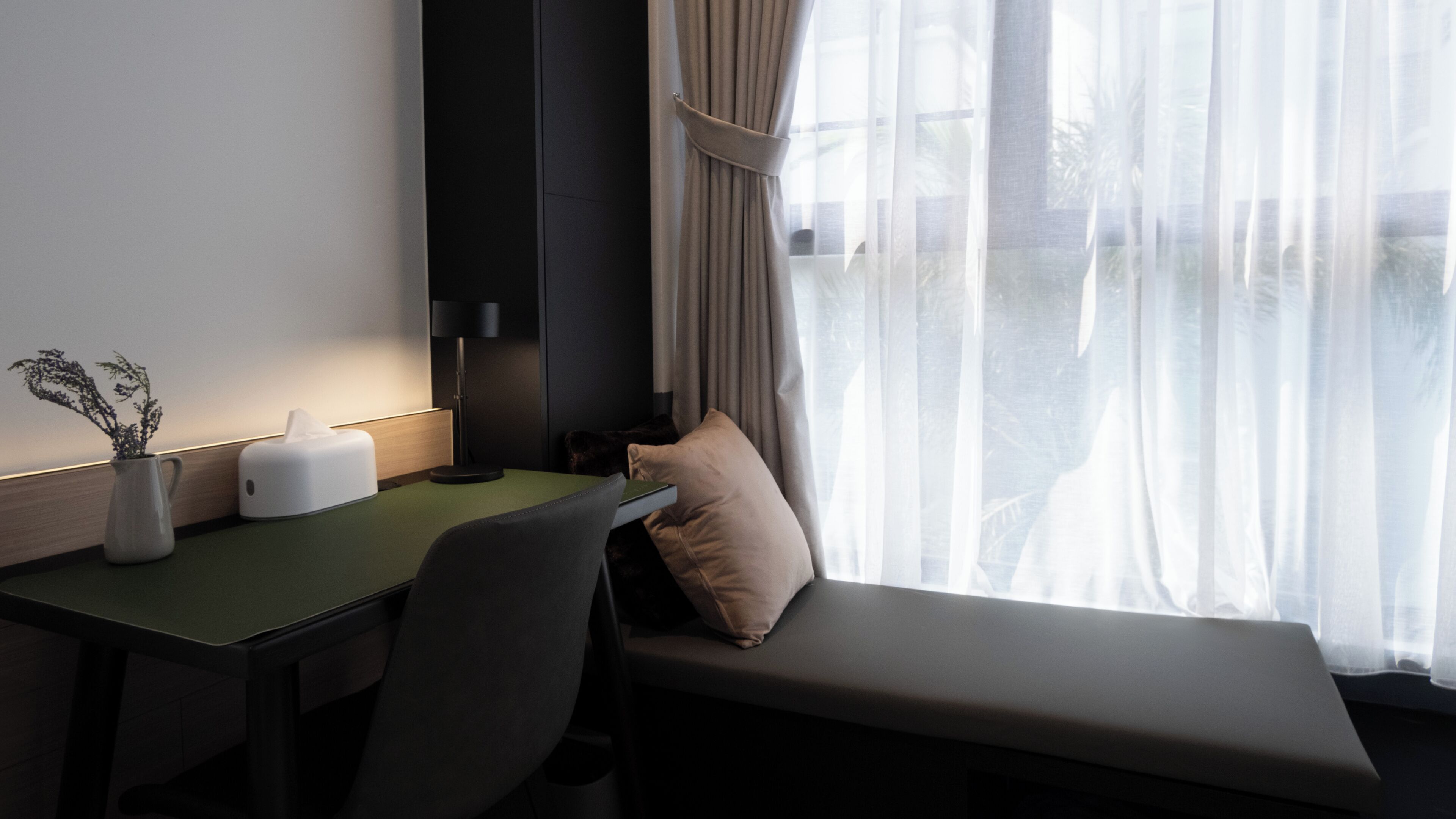 A hotel room with a desk, chair, bedside table, lamp, and a view through sheer curtains.