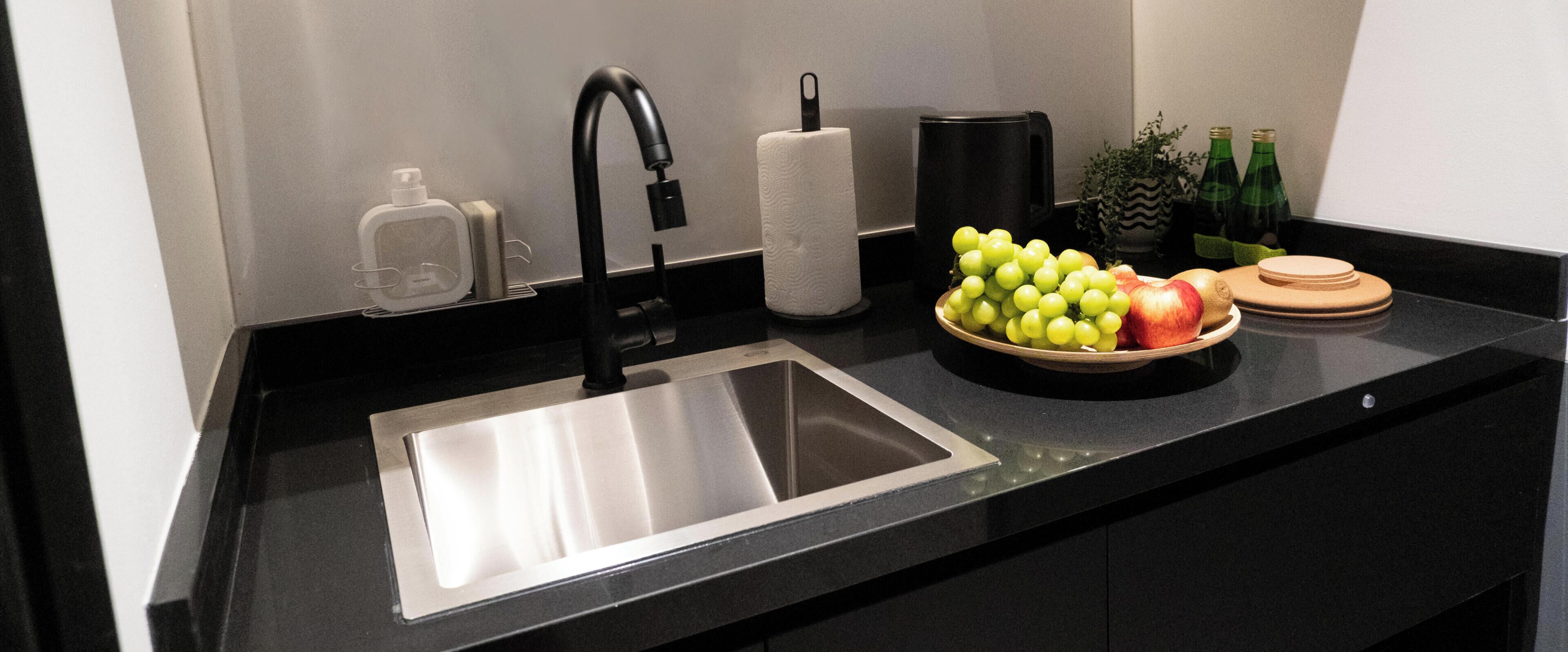 A kitchen with a stainless steel sink, a black faucet, a paper towel holder, a bowl of fruit, and a potted plant.