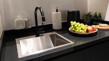 A kitchen with a stainless steel sink, a black faucet, a paper towel holder, a bowl of fruit, and a potted plant.