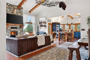 A cozy living room with a stone fireplace, a leather sofa, and a dining area with wooden furniture. - Heated Pool + Hot Tub + Fire Pit + Game Room | Mins to Lake Arrowhead + Village (Lake Arrowhead)
