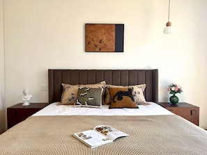 Ein ordentlich bezogenes Bett mit einem gemusterten Überwurf, zwei Kissen mit Vogelmotiven, ein gerahmtes Kunstwerk an der Wand, eine Nachttischlampe und ein Blumenvasen auf dem Nachttisch.