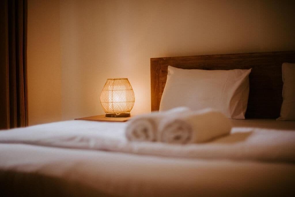 A neatly made bed with rolled towels, a wooden headboard, and a wall-mounted lamp.