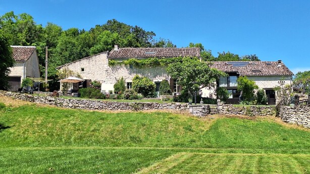 Un bâtiment en pierre, surmonté d’un toit en tuiles, entouré d’un mur de pierre et d’un champ herbeux.
