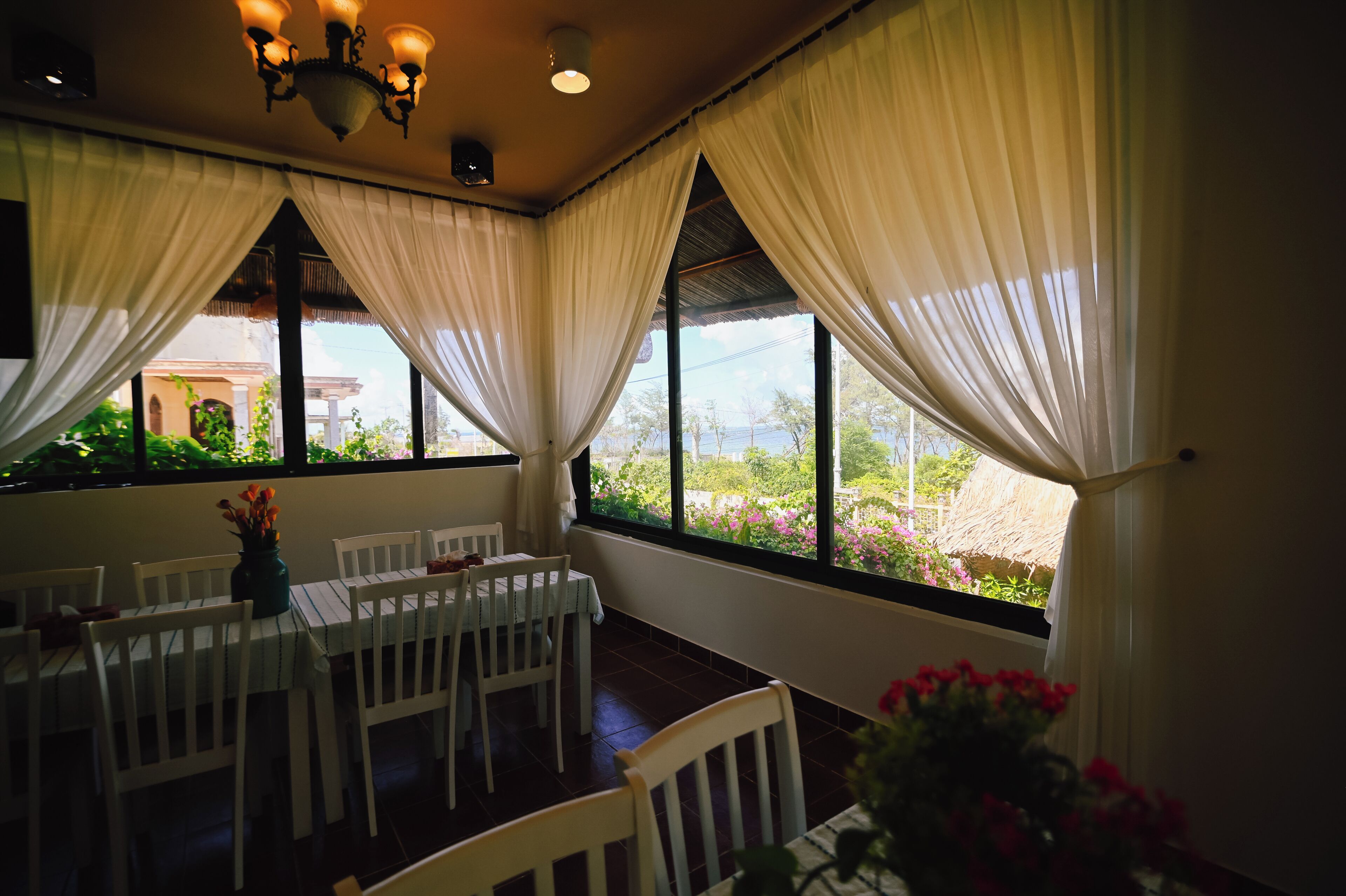 A room with a dining table, chairs, a chandelier, and large windows with curtains.