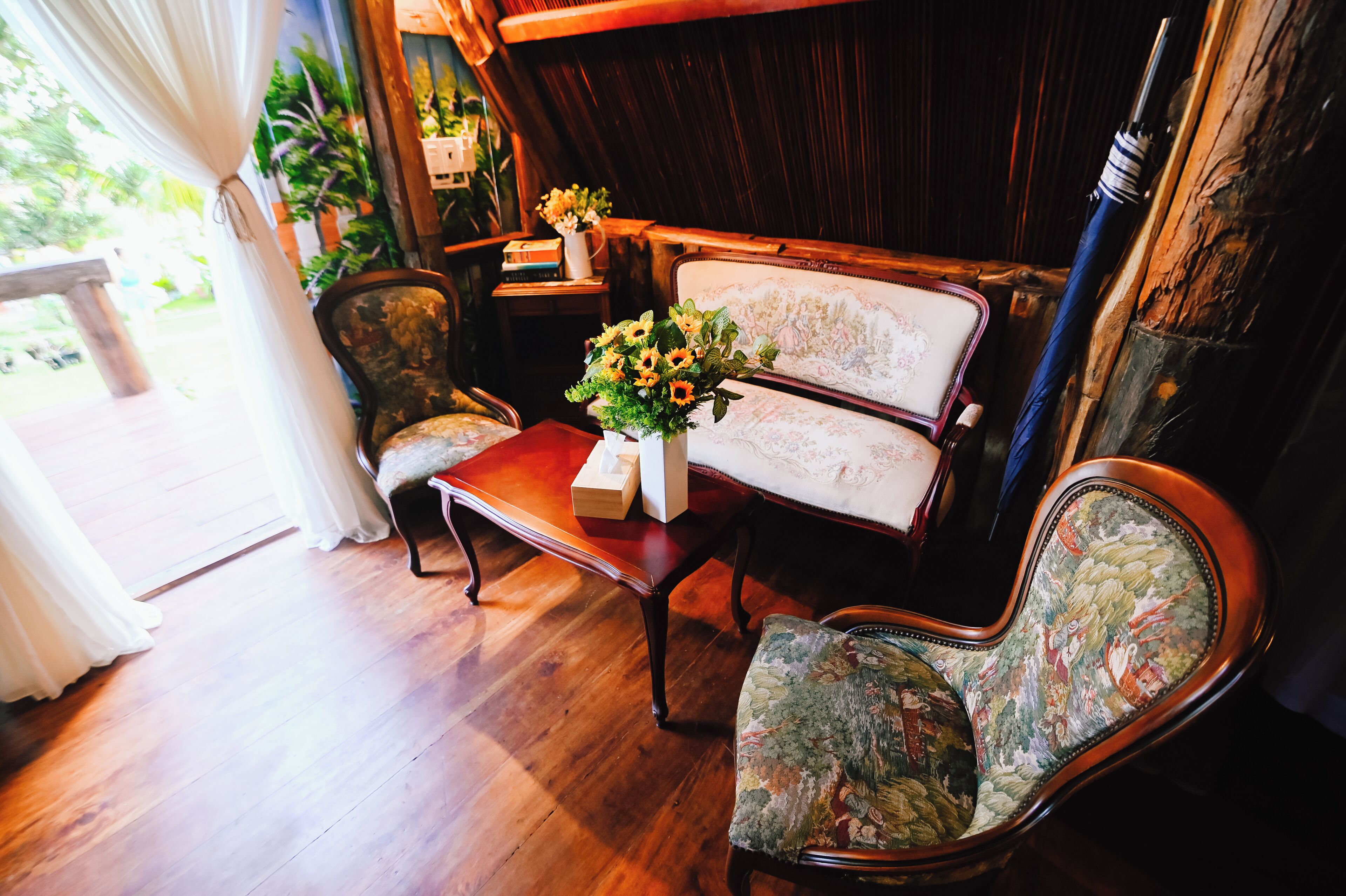 A wooden floor, a sofa with floral upholstery, a table with a vase of flowers, and a chair with a floral pattern.