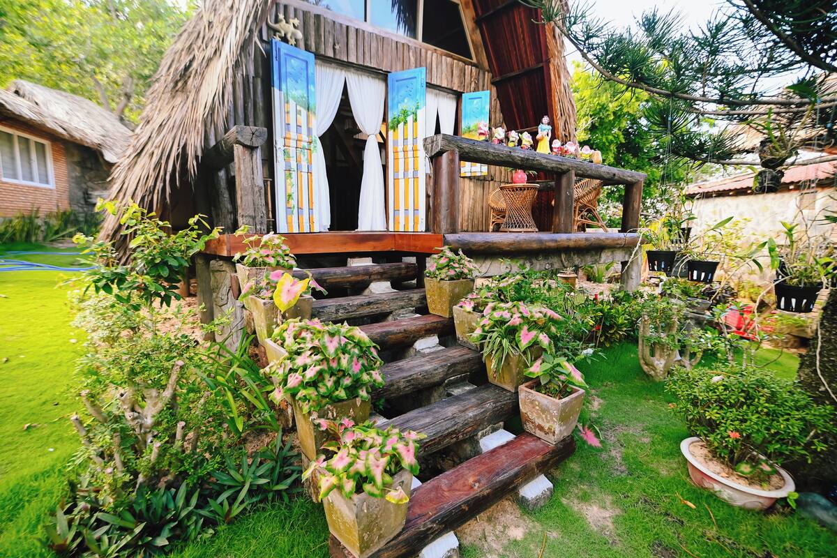 Une maison rustique en bois, avec un toit de chaume, un escalier en bois menant à un porche et des plantes en pot.