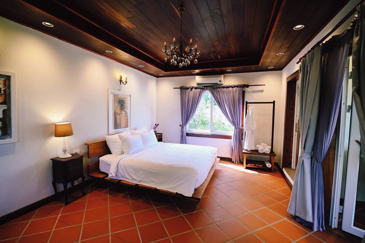 Une chambre à coucher avec un plafond en bois, un lit, une table de chevet, une lampe, une fenêtre avec des rideaux et une porte.
