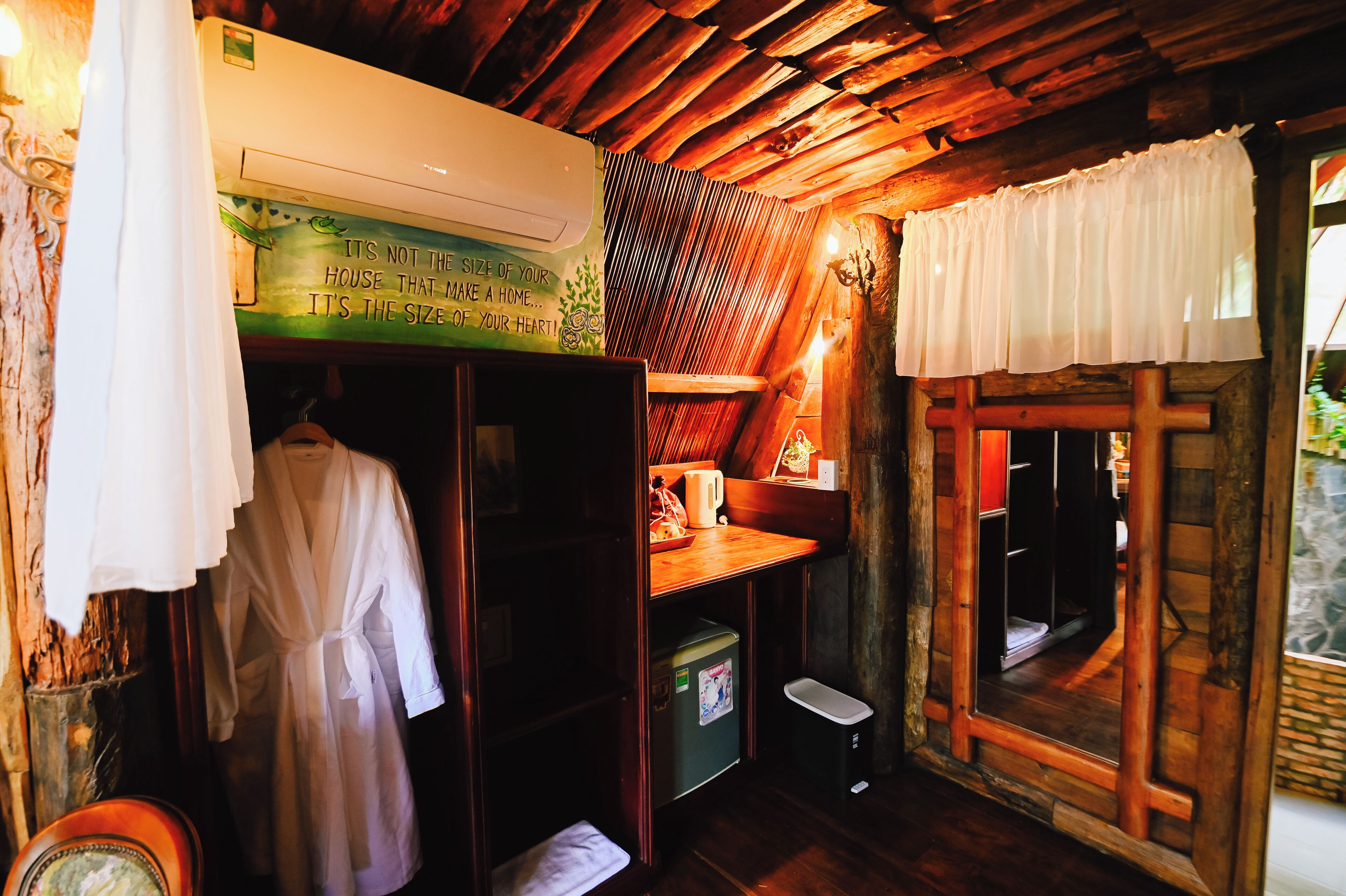 A rustic cabin interior with a wooden ceiling, a small kitchenette, a white robe hanging, and a window with a curtain.